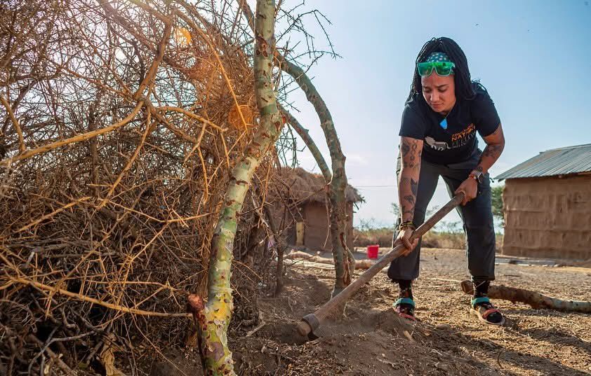 Taylor digging with a hoe beside a fence of thorn branches